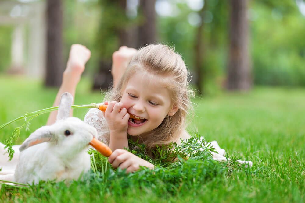Imagem de uma menina loira deitada na grama com um coelho branco. Ela está dando cenouras ao coelho.