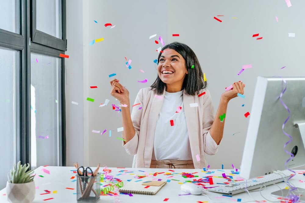 Imagem de uma mulher em seu ambiente de trabalho. Ela está jogando vários confetes para cima, em clima de Natal.