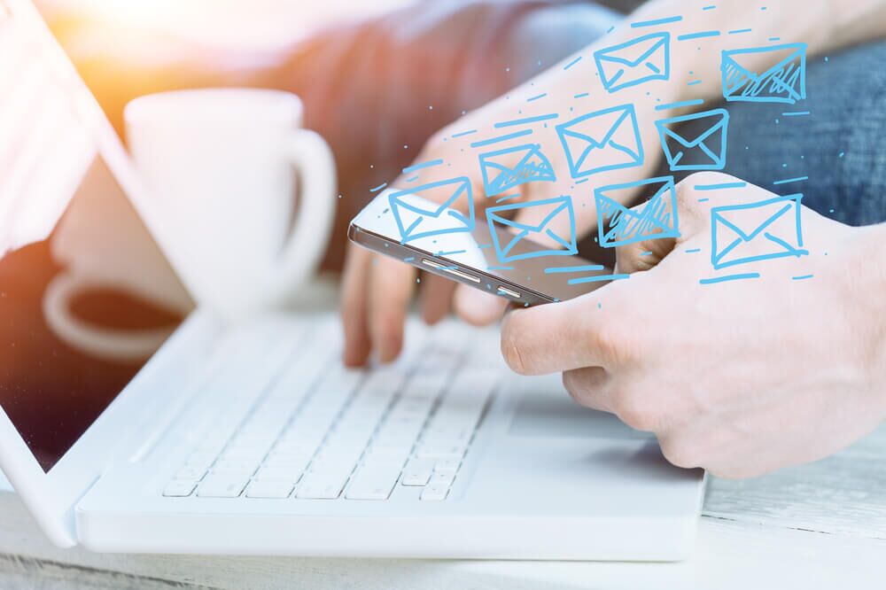 Pessoa digitando no teclado ao mesmo tempo que utiliza o celular. Há desenhos de envelopes de cartas representando as newsletters em azul.