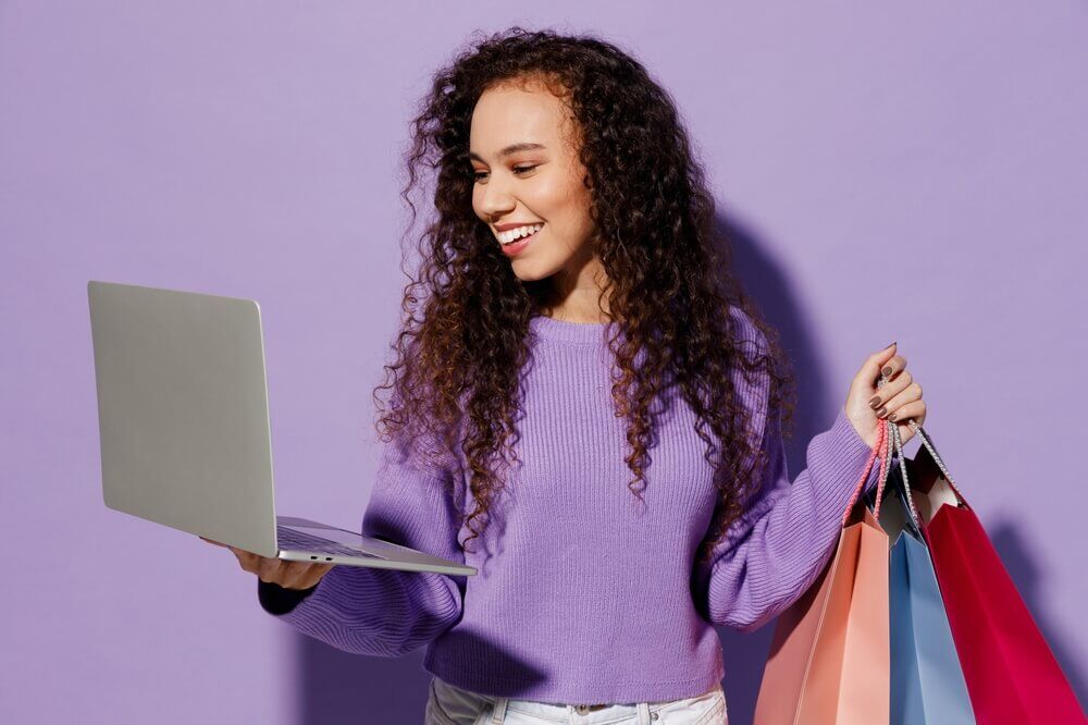 Imagem de uma mulher morena, de cabelos cacheados, vestindo um suéter roxo. Em uma das mãos, elas está segurando sacolas, na outra, um notebook.