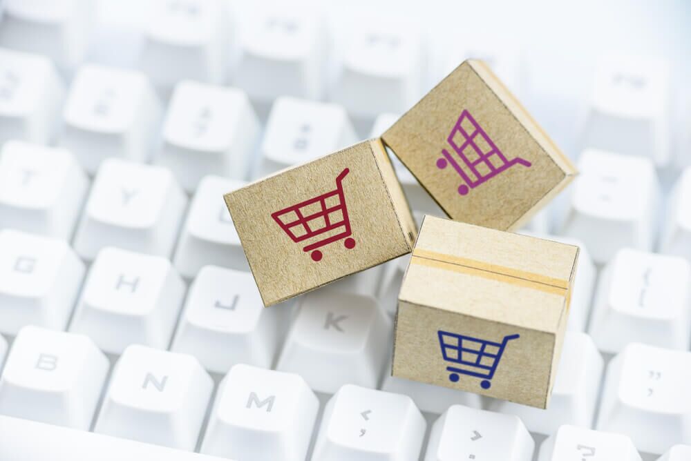 Imagem de pequenas caixinhas com o símbolo de um carrinho, representando compras em um marketplace. Elas estão em cima do teclado de um computador.