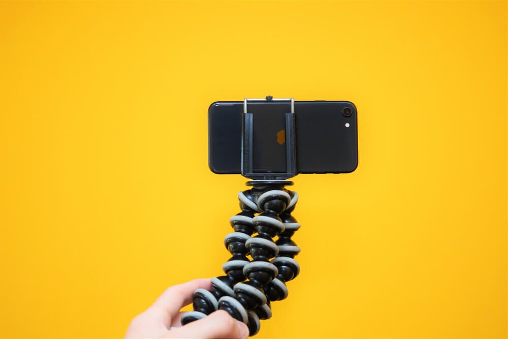 Imagem deu ma mão segurando um tripé com um celular filmando. Ao fundo, é possível notar uma parede amarela.