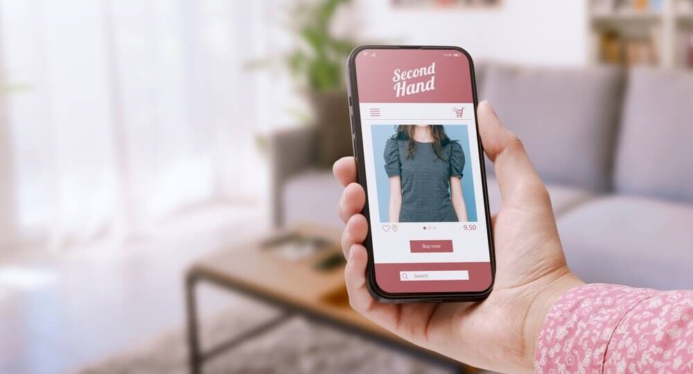Imagem De Uma Pessoa Segurando Um Celular Na Mão. Ela Está Vendo Um Site De Recommerce De Roupas Utilizadas.