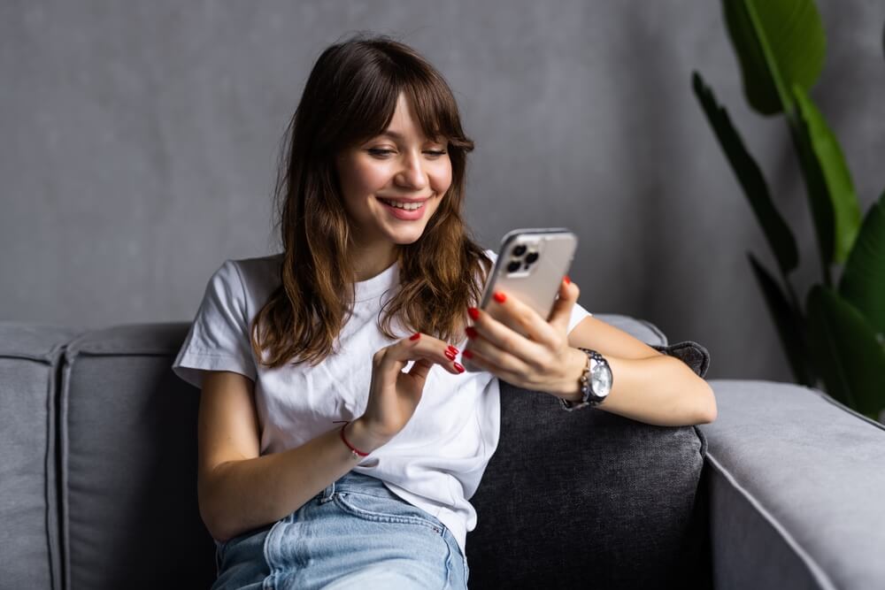 Mulher branca sentada em um sofá. Ela tem franja e os cabelos claros. Está mexendo no celular e sorrindo. Tem relógio nos punhos e esmalte vermelho.